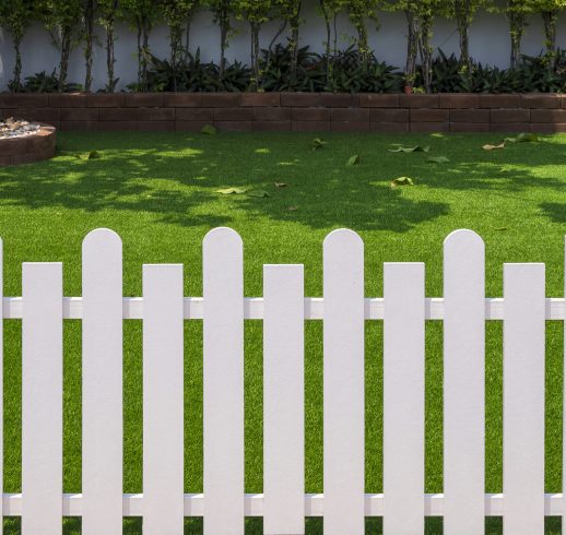 Front view of white wooden fence in front of artificial turf with green plant growing on interlocking brick blocks in front yard of home